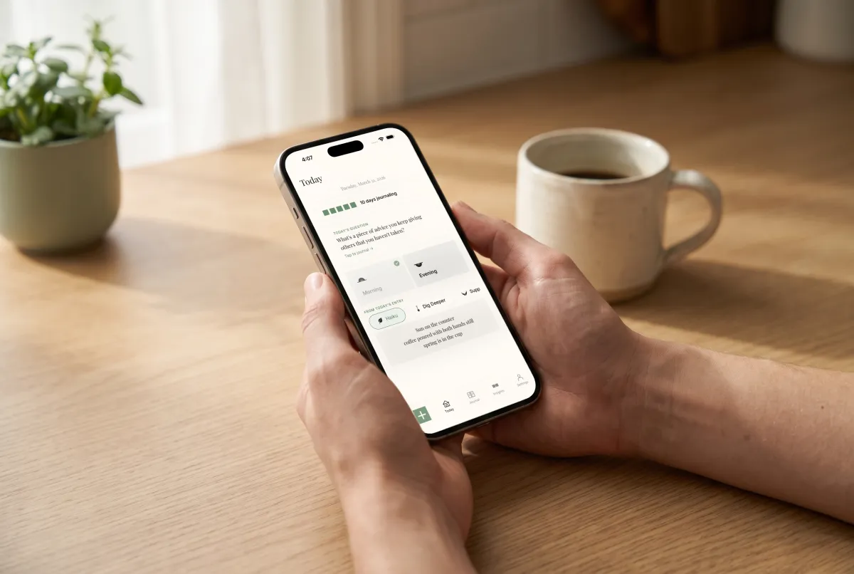Hands holding an iPhone open to the Already There Today screen, with a cup of coffee on the kitchen counter